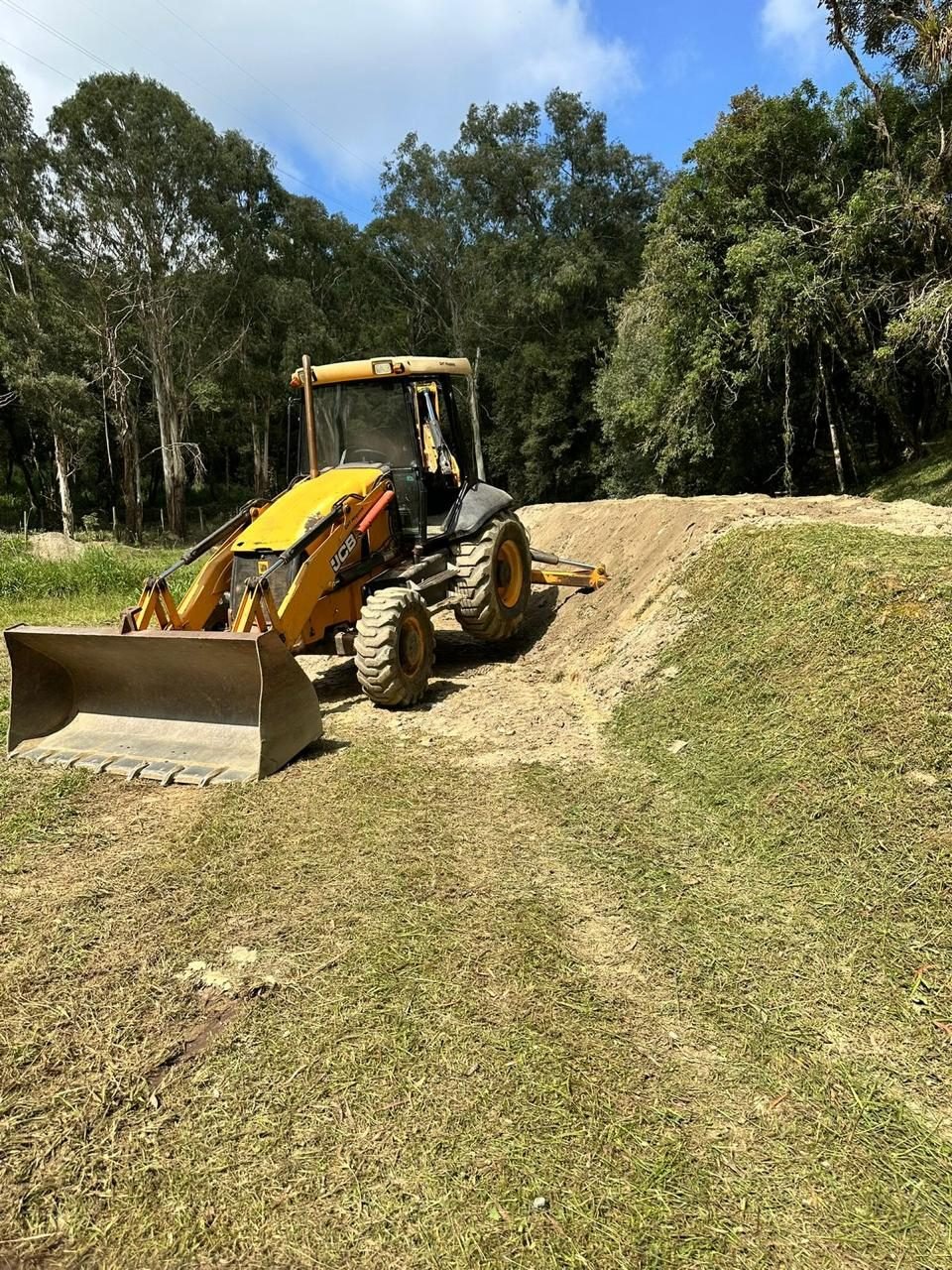 Retroescavadeira JCB realizando nivelamento de terreno em área arborizada